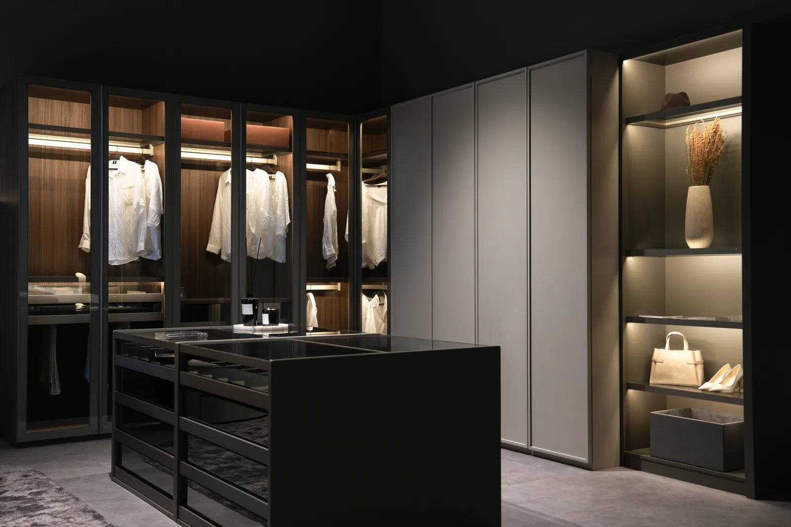 A Lazzoni walk-in closet with a dark glass-topped center island of drawers, reachable from both sides, flanked by glass-front wardrobe units with backlit hanging space and open display shelving holding a vase, handbag, and heels.