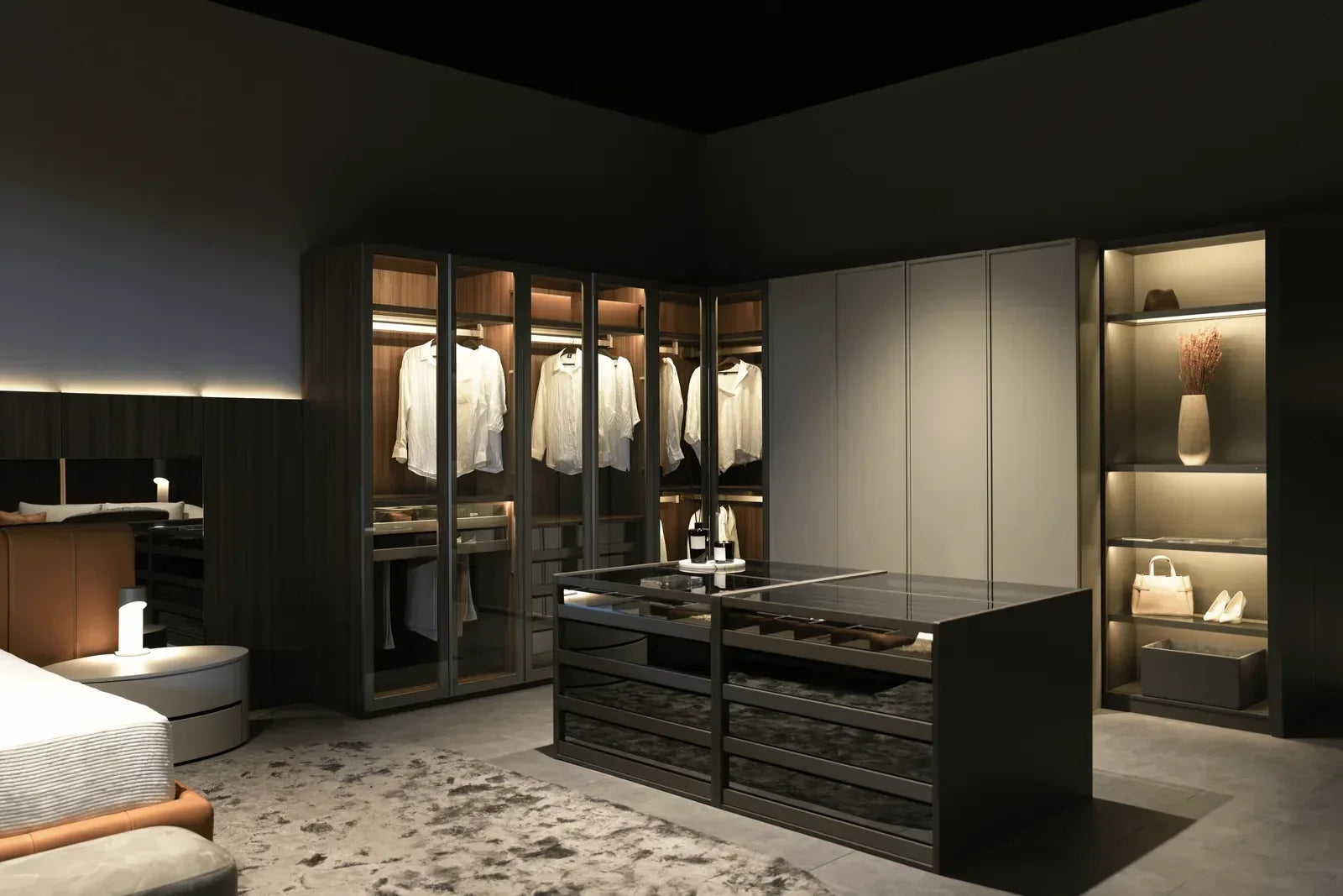 LAZZONI walk-in closet with backlit wood-tone wardrobe cabinets, glass-front shelving displaying a handbag and heels, an upholstered leather bench, and a mirrored center island with drawer storage, set beside a bed with a round nightstand.
