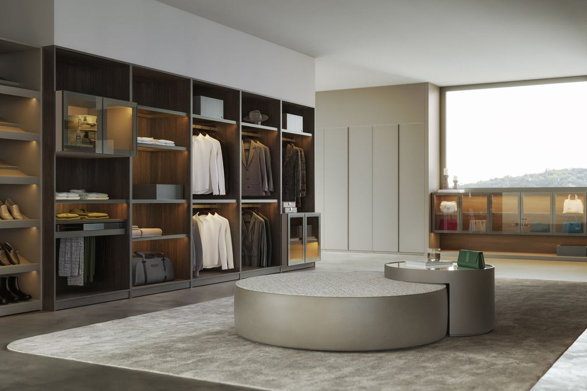 LAZZONI Spazio walk-in closet taking up a whole room, with open walnut shelving and hanging along one wall, a round upholstered bench at the center and daylight from a full-height window