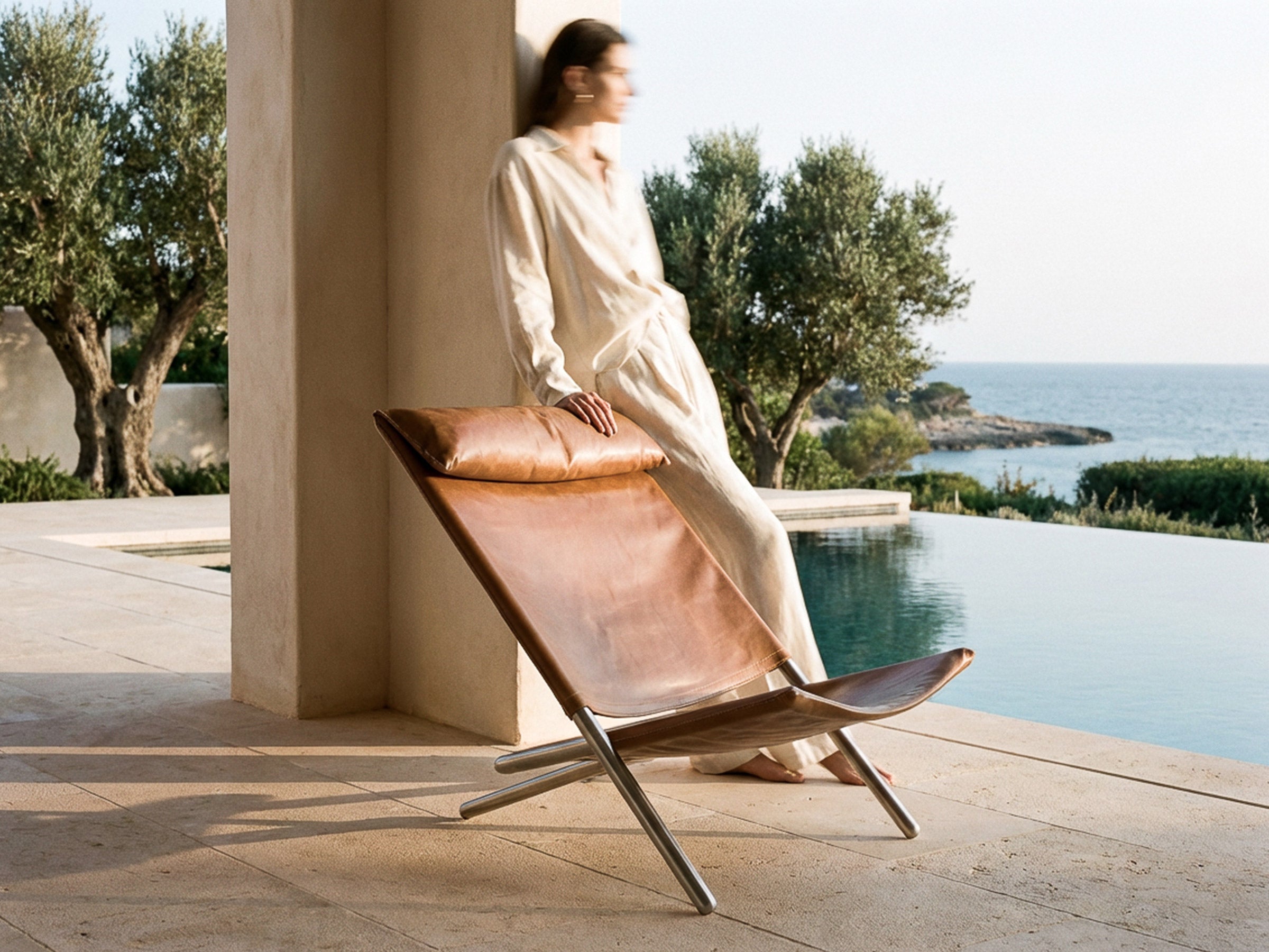 A woman in a cream-colored linen outfit sits on a brown leather lounge chair with a wooden frame, positioned next to an infinity pool with a scenic ocean view in the background.