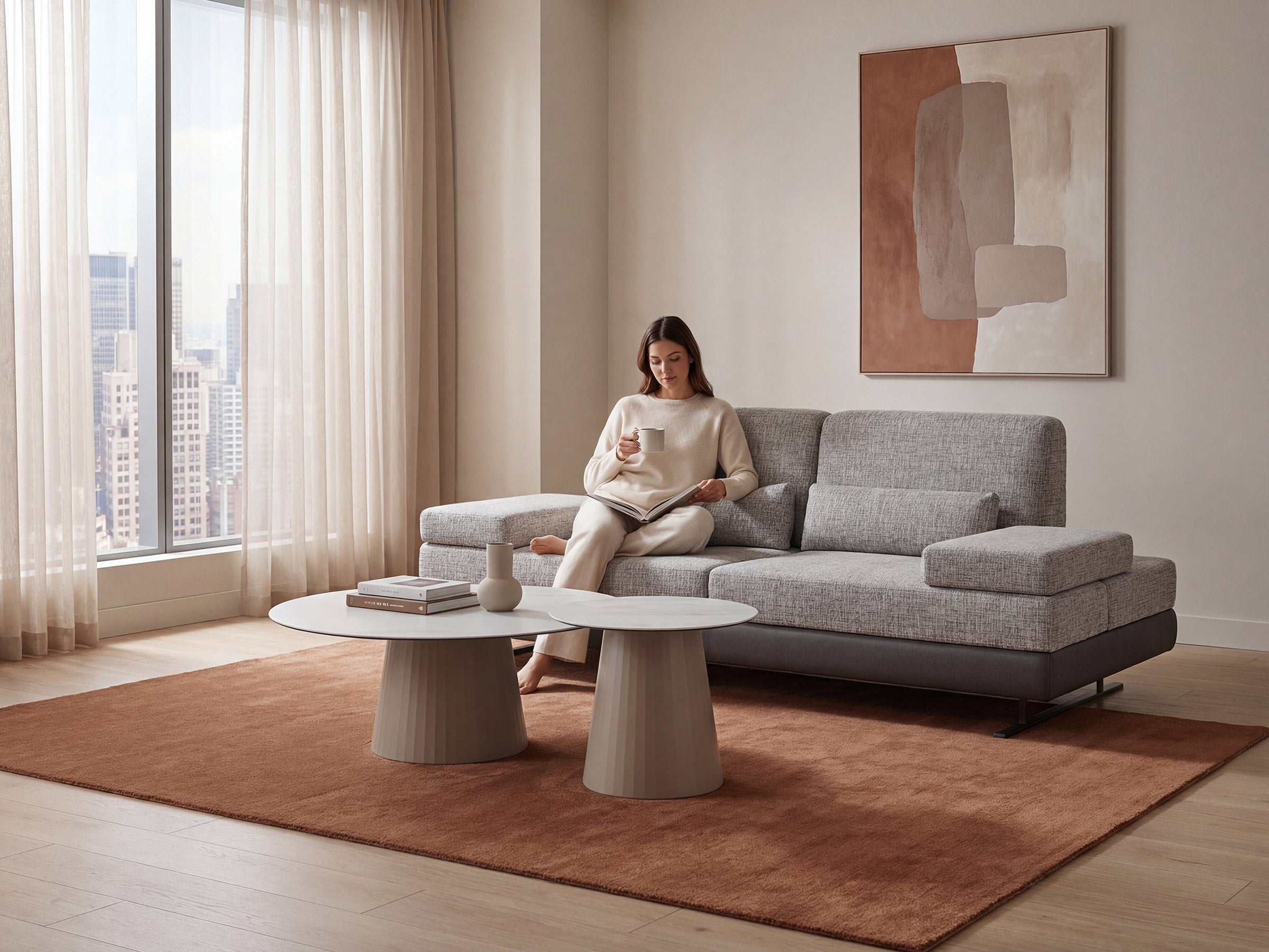 Woman sitting on a gray sofa in a modern living room with large windows and abstract art.